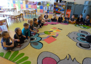 Dzieci siedzą na dywanie wokół rozłożonych figur geometrycznych.